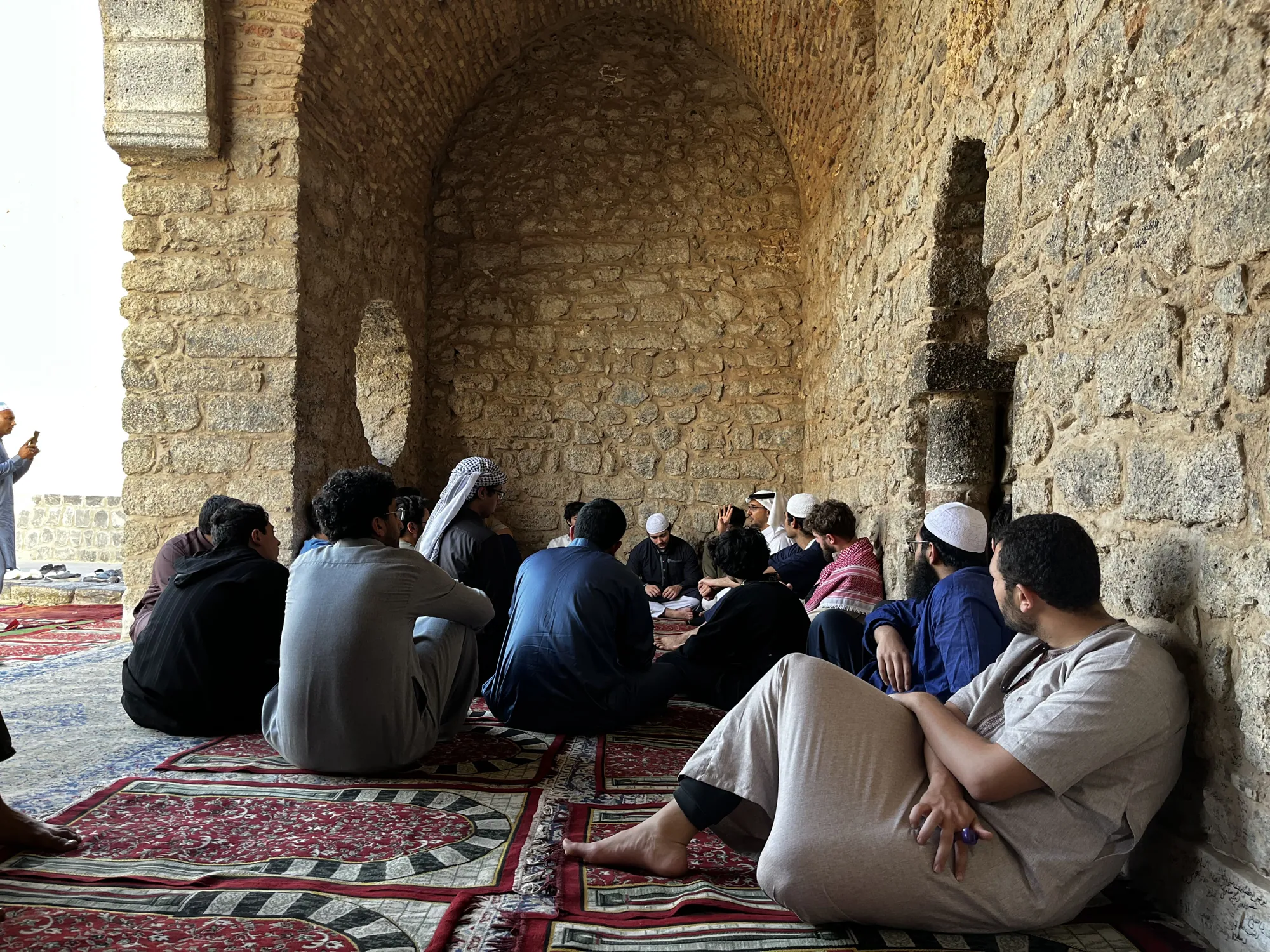 Students seated on patterned rugs inside a historic stone mosque, listening to a teacher during a lesson on Sira history.
