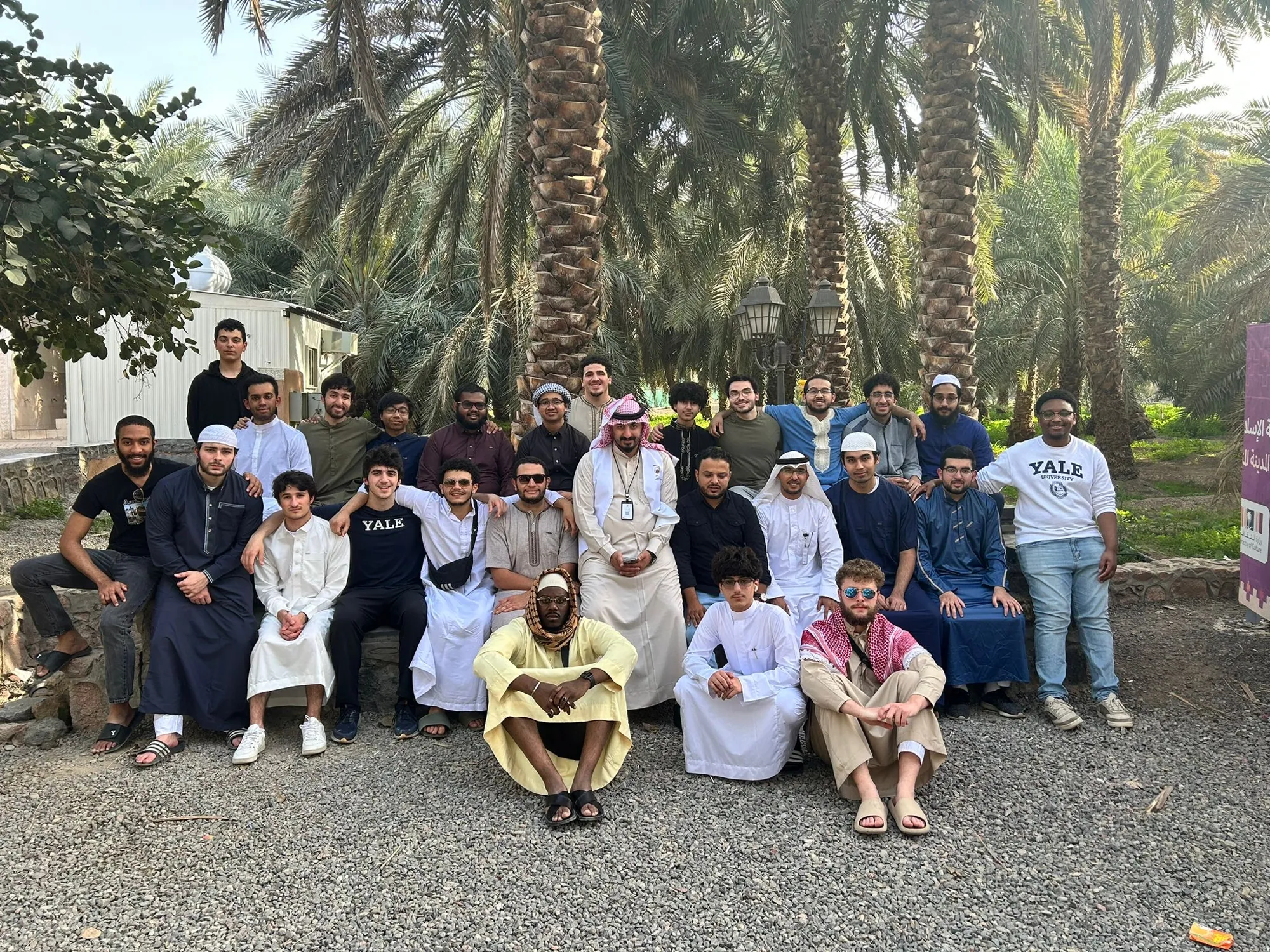 A large group of students standing together beneath date palms during a farm visit in Madinah.